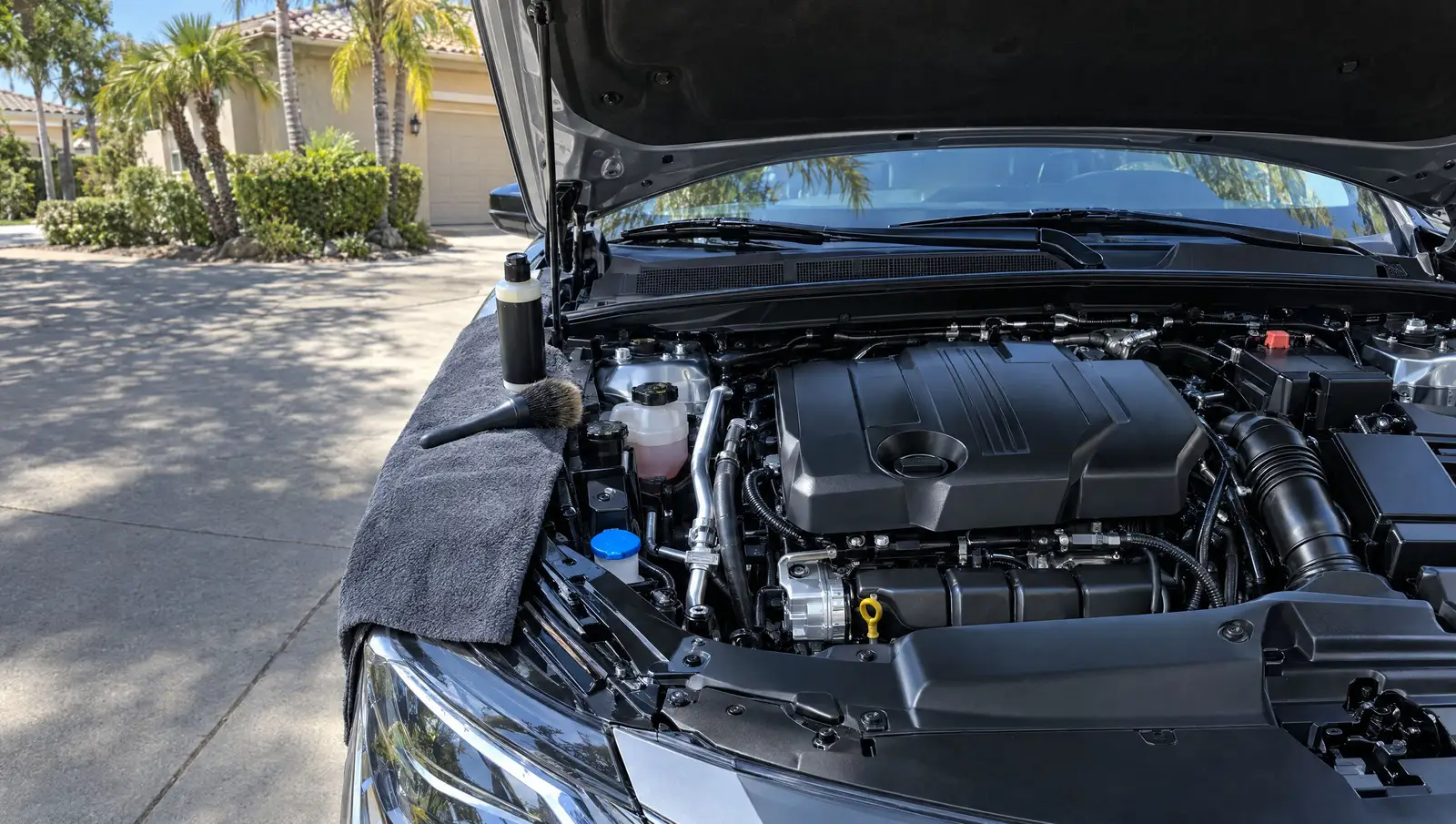 Hood open on a sedan in a driveway, engine bay cleaned and dressed, brush and dressing on the fender