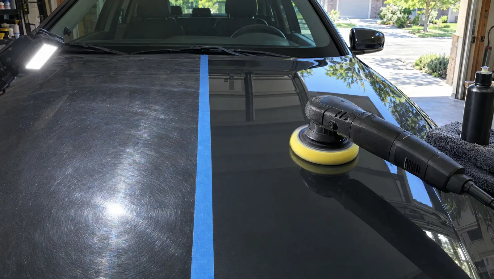 Polisher on a dark hood under an inspection light, tape line between the swirled half and the corrected half